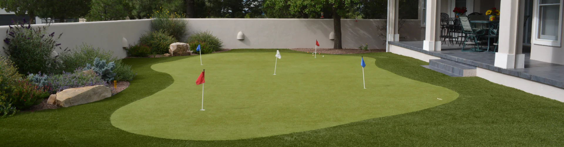 Backyard putting green with red, white, and blue flag pins surrounded by desert garden landscaping, boulder accents, and a covered patio on a stucco-walled residential property